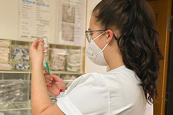 Eine Frau mit Mundschutz steht im Stationszimmer eines Krankenhauses und zieht ein Medikament in eine Spritze.