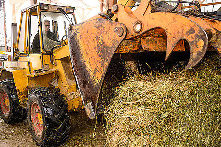 Ein junger Mann sitzt am Steuer eines orangefarbenen Traktors und lenkt die Greifschaufel auf einen großen Heuballen.