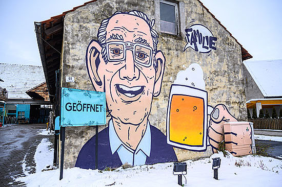 Eine Hauswand mit großer Graffiti-Zeichnung: Ein älterer Mann hält eine Maß Bier, darüber eine Sprechblase mit dem Wort: „Familie“. Daneben steht ein Schild mit der Aufschrift: „Geöffnet“.   