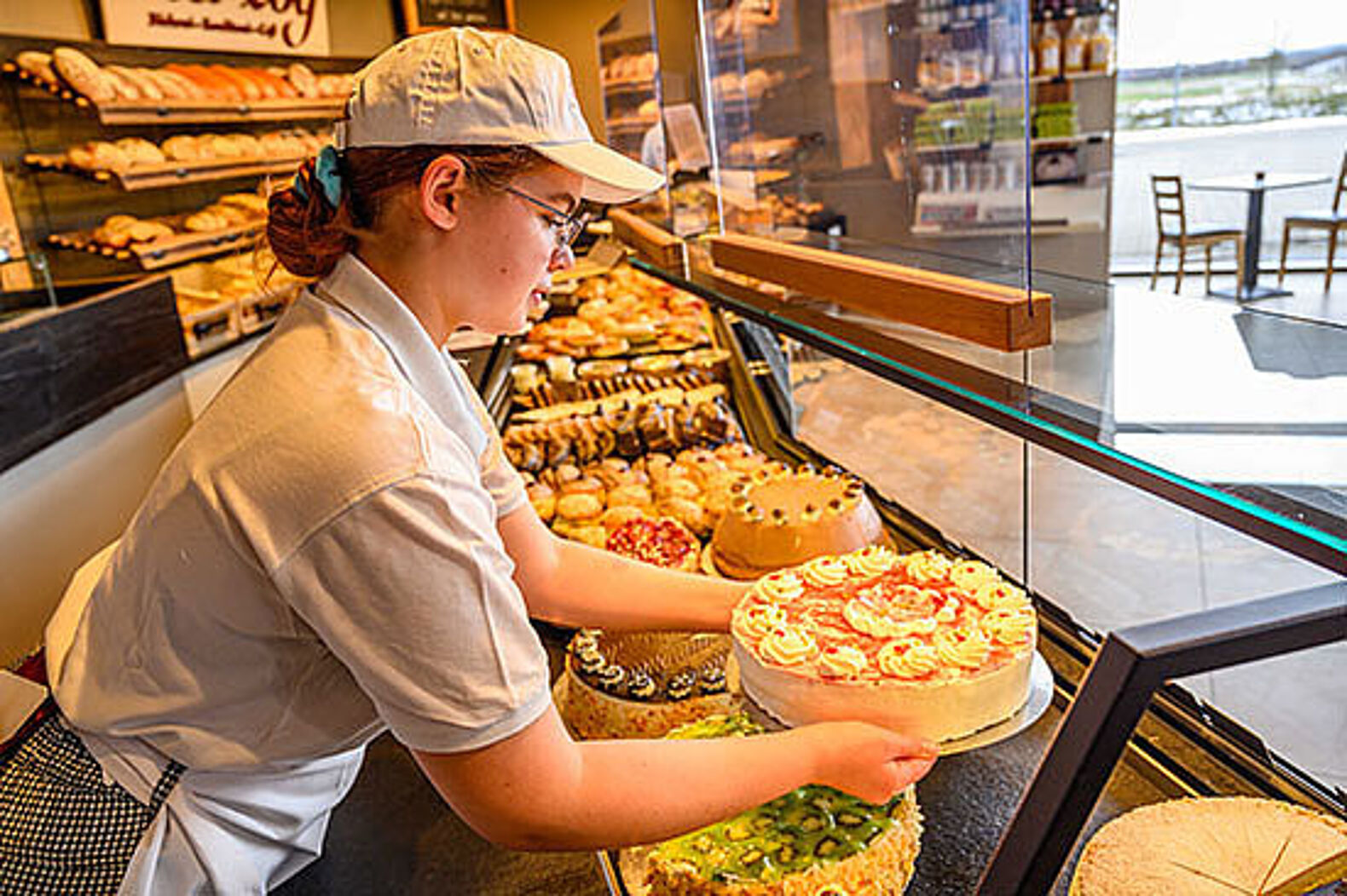 Eine junge Frau mit Schürze stellt eine Torte zu anderen Torten in eine Café-Auslage.