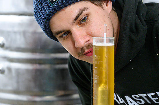 Ein junger Mann schaut auf ein Reagenzglas mit gelb-brauner Flüssigkeit. Darin steckt eine Spindel. 