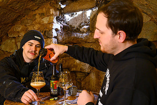 Zwei Männer sitzen an einem Tisch in einem Gewölbekeller. Einer gießt dem anderen Bier in sein Glas. 