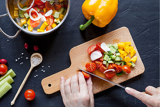 Hand mit Messer schneidet Gemüse auf Schneidebrett, gelbe Paprika und Schüssel daneben.