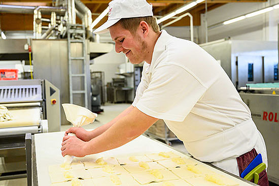 Ein junger Mann in Bäckerkleidung drückt eine Masse aus einer Spritztüte auf Teigquadrate auf einem Tisch. 