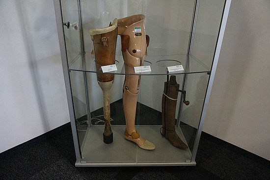 Alternativtext	Detailaufnahme: Gipsabdruckhüllen von drei Füßen auf einer Werkbank 	Detailaufnahme: Glasvitrine mit drei historischen Beinprothesen