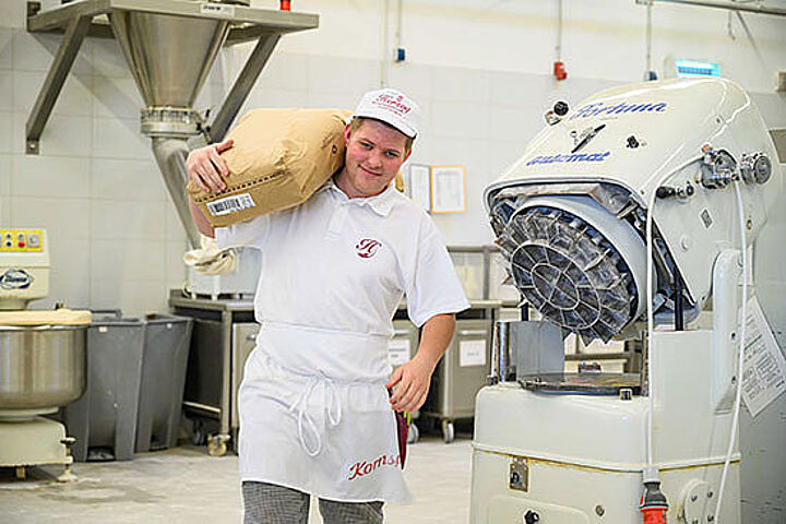 Ein junger Mann mit Schürze und Cap trägt einen Sack auf der Schulter durch eine Halle mit Maschinen. 