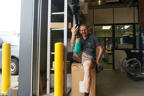 Matthias sitzt auf einer Kiste und streckt seine Beinprothese senkrecht in die Höhe. 