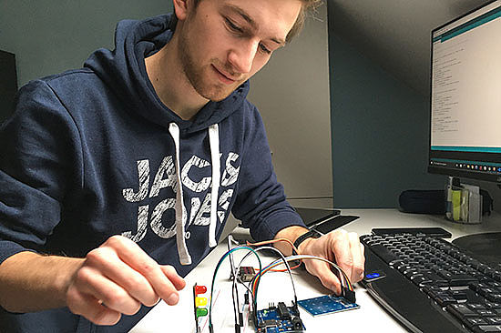 Ein Mann sitzt an einem Tisch und zeigt einen sogenannten Mikro-Controller mit mehreren Kabelverschaltungen. 