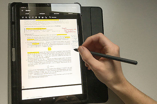 Ein Tablet-Bildschirm zeigt Text und mathematische Formeln. Daneben eine Hand mit elektronischem Stift.  