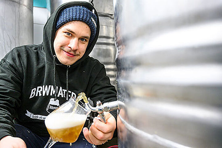 Ein junger Mann mit Mütze und Kapuzenpulli zapft ein Glas Bier aus einem großen Aluminium-Fass. 