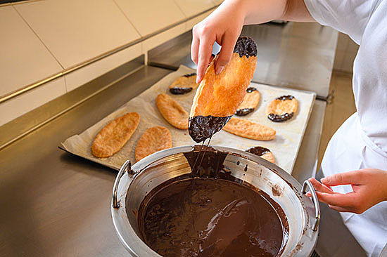 Eine Hand zieht ein ovales Gebäckstück aus einer Schüssel mit flüssiger Schokolade. 